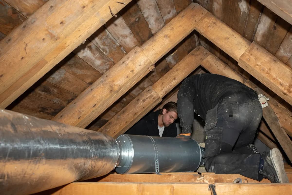 Två tekniker i arbetskläder monterar en stor isolerad ventilationskanal mellan takstolarna på en vind