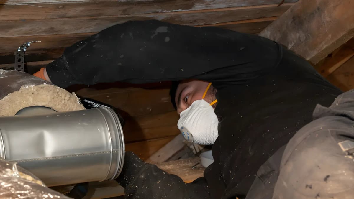 Ventilationstekniker med andningsskydd ligger på sidan i ett trångt vindsutrymme i Stockholm intill en isolerad kanal
