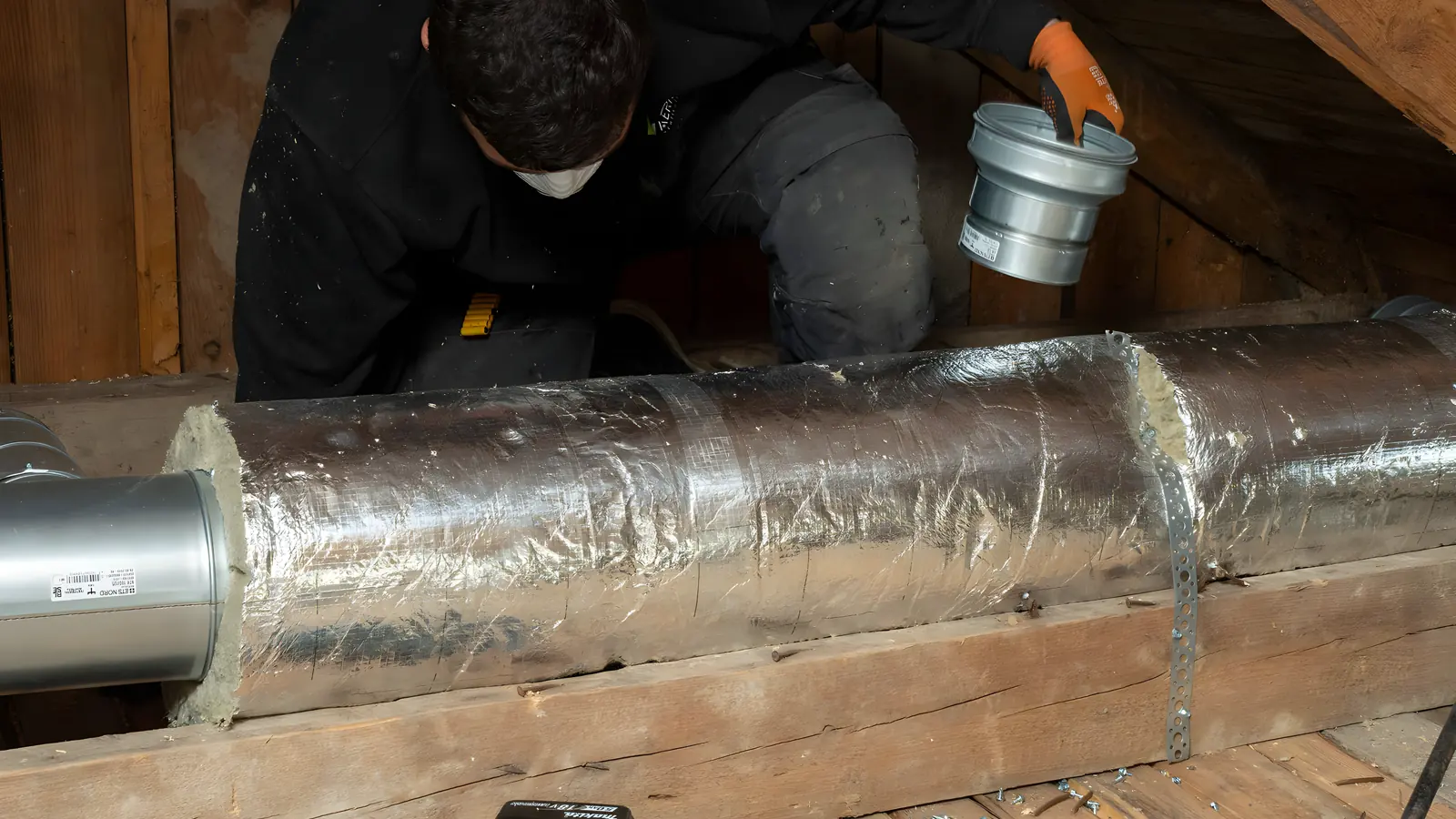 Montör på knä under vindstaket håller upp en blank plåtkoppling ovanför en isolerad ventilationskanal mellan bjälkarna
